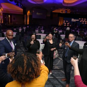 The image shows a group of people standing in a circle, raising glasses in a toast. They are in a large, dimly lit ballroom with round tables and chairs arranged throughout the room. The lighting is a mix of warm and purple hues, highlighting the ceiling's arches and creating a festive atmosphere. In the background, there is a stage with a large screen displaying the text 
