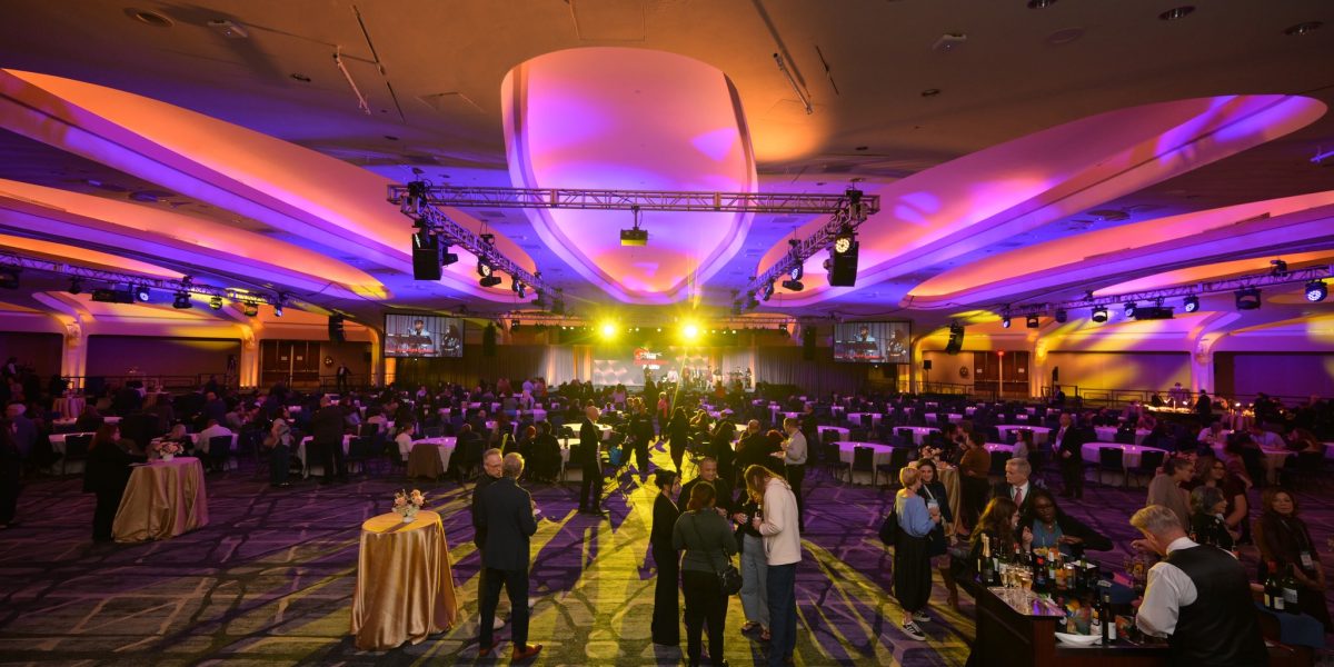 The image depicts a large, indoor event space filled with people. The ceiling is high and features modern architectural lighting with shades of purple and orange, casting vivid colors across the room. The venue is spacious with rows of tables, each draped with tablecloths and hosting guests seated in groups. In the foreground, several people stand in small clusters, engaging in conversation near high tables adorned with small floral arrangements. To the right, a bartender is serving drinks at a well-stocked bar. The background displays a stage with a bright yellow spotlight, where several screens show images of the event. The overall atmosphere is lively and vibrant.