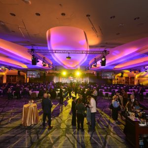 The image depicts a large, indoor event space filled with people. The ceiling is high and features modern architectural lighting with shades of purple and orange, casting vivid colors across the room. The venue is spacious with rows of tables, each draped with tablecloths and hosting guests seated in groups. In the foreground, several people stand in small clusters, engaging in conversation near high tables adorned with small floral arrangements. To the right, a bartender is serving drinks at a well-stocked bar. The background displays a stage with a bright yellow spotlight, where several screens show images of the event. The overall atmosphere is lively and vibrant.