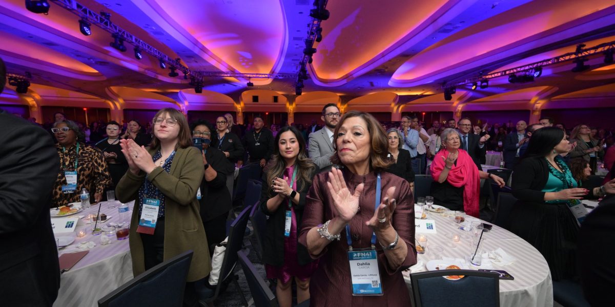 The image depicts a large group of people standing and clapping in a spacious conference hall. The ceiling is illuminated with vibrant blue and purple lighting, creating an energetic atmosphere. Round tables with white tablecloths are scattered throughout the room, some with empty plates and glasses visible. Attendees, wearing conference badges, are of diverse ages and are dressed in professional attire. In the foreground, a woman in a burgundy dress applauds. Behind her, several individuals are also clapping, indicating a moment of celebration or recognition.