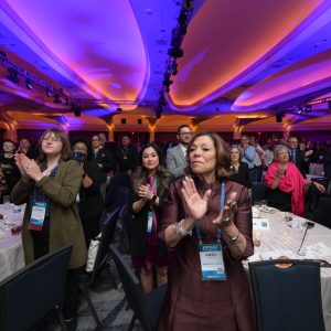 The image depicts a large group of people standing and clapping in a spacious conference hall. The ceiling is illuminated with vibrant blue and purple lighting, creating an energetic atmosphere. Round tables with white tablecloths are scattered throughout the room, some with empty plates and glasses visible. Attendees, wearing conference badges, are of diverse ages and are dressed in professional attire. In the foreground, a woman in a burgundy dress applauds. Behind her, several individuals are also clapping, indicating a moment of celebration or recognition.
