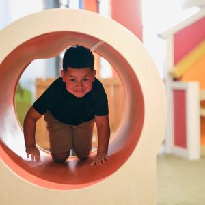 Child crawling through a wooden tunnel in a colorful indoor play area.