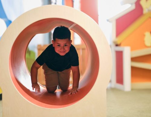 Child crawling through a wooden tunnel in a colorful indoor play area.
