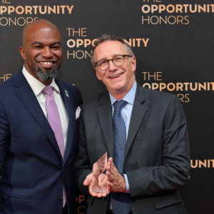 Two men in suits standing together, one holding an award, against a backdrop reading "THE OPPORTUNITY HONORS."