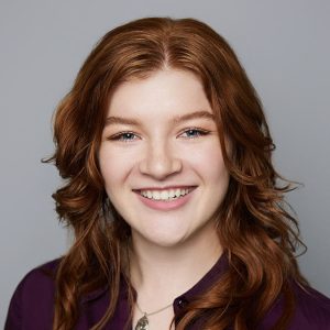 A white person with long red hair is facing the camera and smiling. They are wearing a purple top and a silver necklace. The background is gray. - Jordan Kennedy-Rea