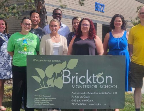 A group of nine adults standing outside a building with yellow bricks, behind a green sign that says, "Brickton Montessori School."