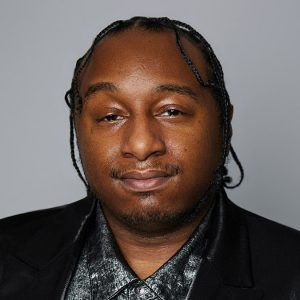 A Black man with braided hair is facing the camera. He is wearing a gray shirt, a black blazer, and a silver necklace. The background is gray. - Joshua Maratre