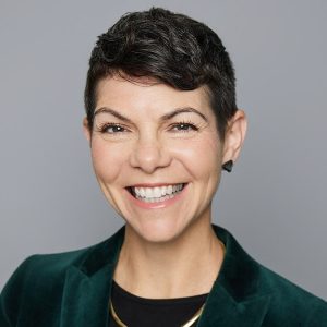 A white woman with short brown hair is facing the camera and smiling. She is wearing a black top, with an emerald blazer, a gold necklace, and black earrings. The background is gray. - Mollie Dowling