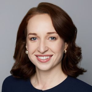 A white woman with long red hair is facing the camera and smiling. She is wearing a navy top and pearl earrings. The background is gray. - Molly Smith
