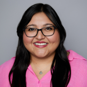 A Latine woman with long straight black hair is facing the camera and smiling. She is wearing a pink shirt and glasses. The background is gray. - Raquel Venado