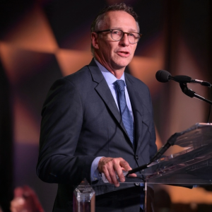 A man in a suit and tie speaks at a transparent podium with a microphone in a dimly lit room with a geometric patterned background.