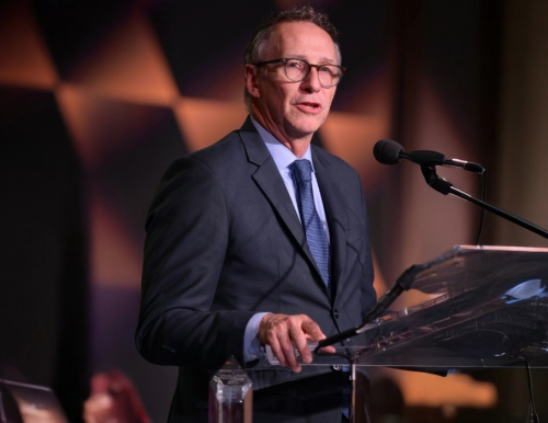 A man in a suit and tie speaks at a transparent podium with a microphone in a dimly lit room with a geometric patterned background.