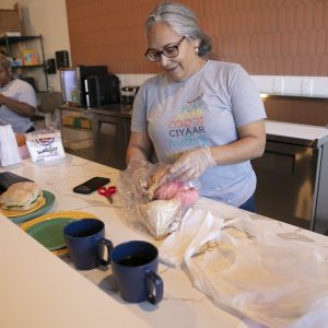 A woman arranging items on a kitchen counter with mugs, a sandwich, and utensils around her.