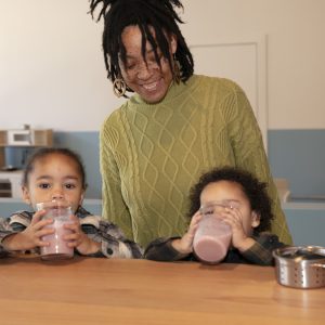 A woman with dreadlocks smiles at two children drinking from glasses at a wooden counter.