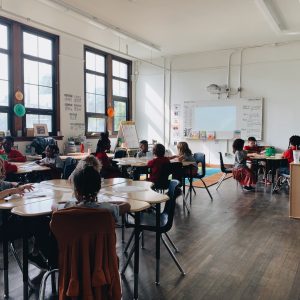 Students seated at round tables in a sunlit classroom.