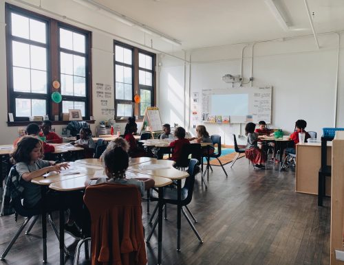 Students seated at round tables in a sunlit classroom.