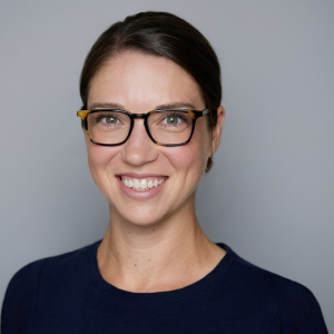 A white woman with brown hair pulled back is facing the camera and smiling. She is wearing a blue shirt and glasses. The background is gray. - Emily Laflamme
