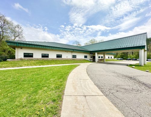 A single-story building with a green metal roof and white exterior sits in a park-like setting. A curving driveway and pathway lead to the entrance. Cloudy sky above.