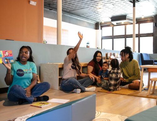 A group of adults and children interacting in an educational indoor setting with a woman holding a book.