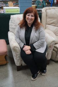 A woman seated on a patterned armchair, wearing a gray blazer and black clothing, in a room with assorted furniture.