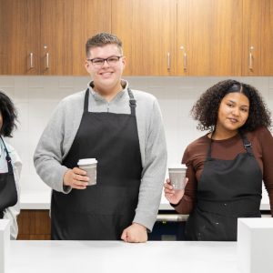 Three people wearing black aprons stand behind a counter holding cups, with wooden cabinets and white tile in the background.