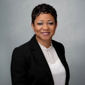 A smiling woman with short dark hair wearing a black blazer over a white blouse. The background is gray.