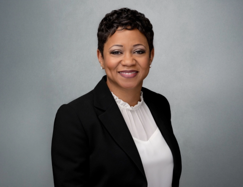 A smiling woman with short dark hair wearing a black blazer over a white blouse. The background is gray.