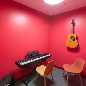 Interior of a small practice room with red walls, an electric keyboard, an acoustic guitar on the wall, and two chairs