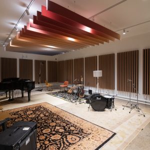 Interior of a recording studio's recording room, with a piano, drum set, xylophone, several chairs, and microphone stands; there are sound dampening panels along the ceiling and walls, and there are rugs on the ground