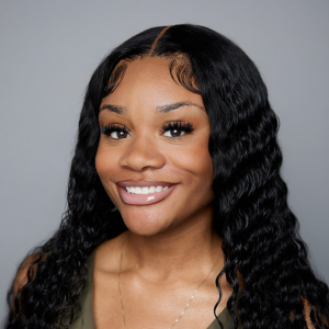 A Black woman with wavy long black hair is facing the camera and smiling. She is wearing a green shirt and a silver necklace. The background is gray. - Kristina Henderson