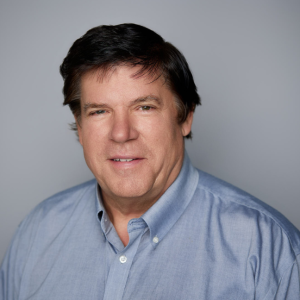 A white man with short black hair is facing the camera and smiling. He is wearing a blue shirt. The background is gray. - Tim Bohnert