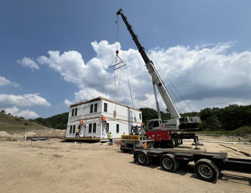 A crane installs a two-story modular building at a construction site with workers guiding it.