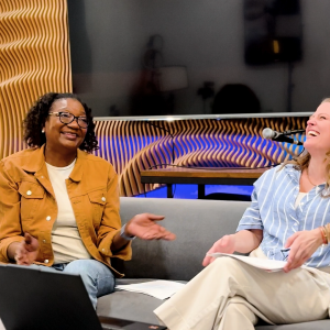 Two women sit together on a sofa laughing in front of a wall with wooden paneling, a TV, and a colorful painting.