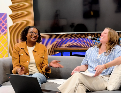 Two women sit together on a sofa laughing in front of a wall with wooden paneling, a TV, and a colorful painting.