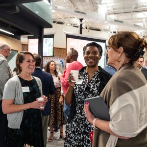 Three women stand inside smiling and talking to each other among a crowd of other people.