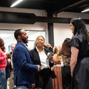 One man and two women stand together and talk, smiling, inside among a group of other people.