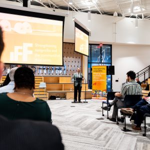 A man stands inside behind a podium and in front of a projection screen and speaks to a group of seated people.