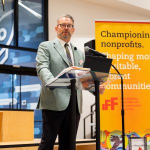 A man stands inside behind a podium speaking into a microphone, in front of a banner that reads, 