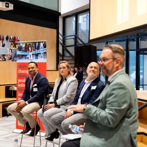 Four people sit inside on tall chairs, three grouped to the left and one to the right, in front of a projection screen.