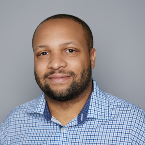 A man with short black hair and facial hair is facing the camera and smiling. He is wearing a blue checkered shirt. The background is gray. - Gabe Guillory