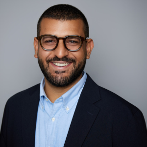 A man with short black hair and facial hair is facing the camera and smiling. He is wearing a black suit jacket and blue shirt. The background is gray. - Yousef Abdo