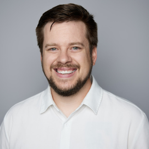 A white man with light brown hair and facial hair is facing the camera and smiling. He is wearing a white shirt. The background is gray. - Zack Daley