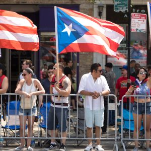 People stand behind a barricade at a parade, holding and waving Puerto Rican flags on a sunny day.