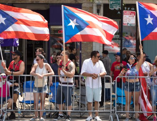 People stand behind a barricade at a parade, holding and waving Puerto Rican flags on a sunny day.