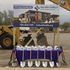 A table covered in a purple table cloth and covered with white construction helmets, which has six shovels leaning on it, sits in front of construction equipment and a sign showing the rendering for a new building