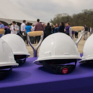 Four white construction helmets on a purple table in front of four shovels and a crowd of people outside