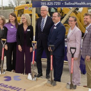 Seven people smile wearing suits or hospital scrubs hold shovels and stand on dirt in front of construction equipment