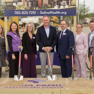 Seven people smile wearing suits or hospital scrubs hold shovels and stand on dirt in front of construction equipment