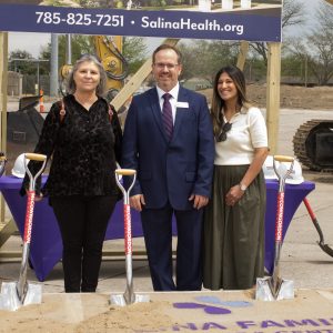 Three people smile wearing suits or hospital scrubs hold shovels and stand on dirt in front of construction equipment