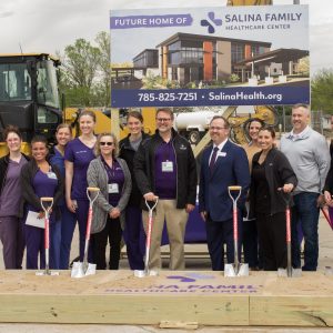 A crowd of people smile wearing suits or hospital scrubs hold shovels and stand on dirt in front of construction equipment